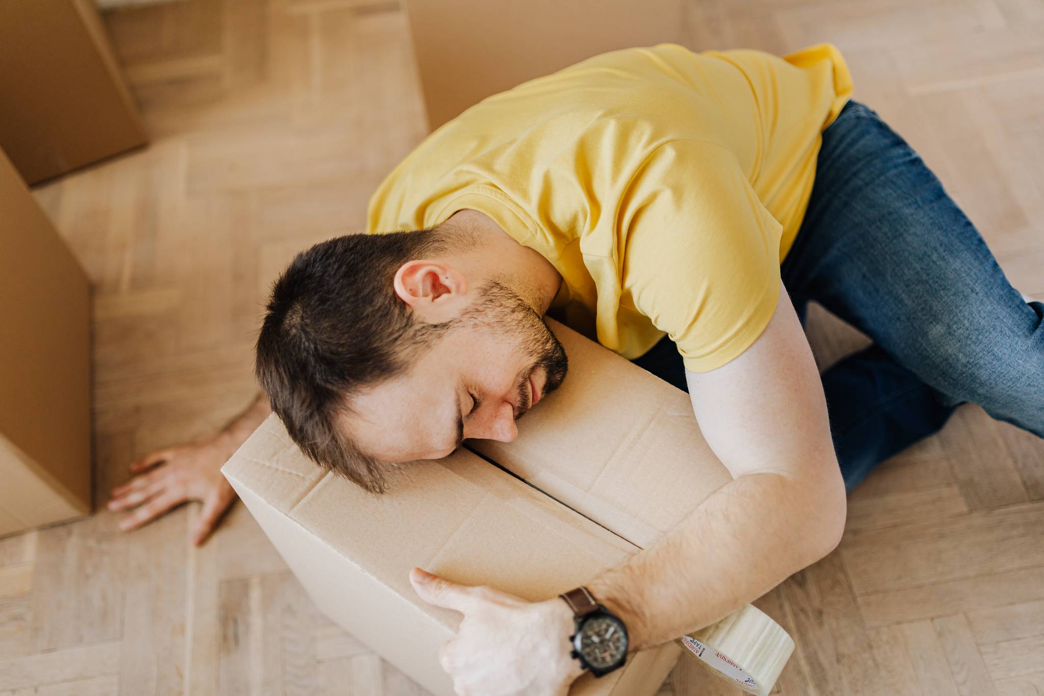 High angle of fatigued male in casual wear sleeping on floor with cardboard box after packing belongings before moving out